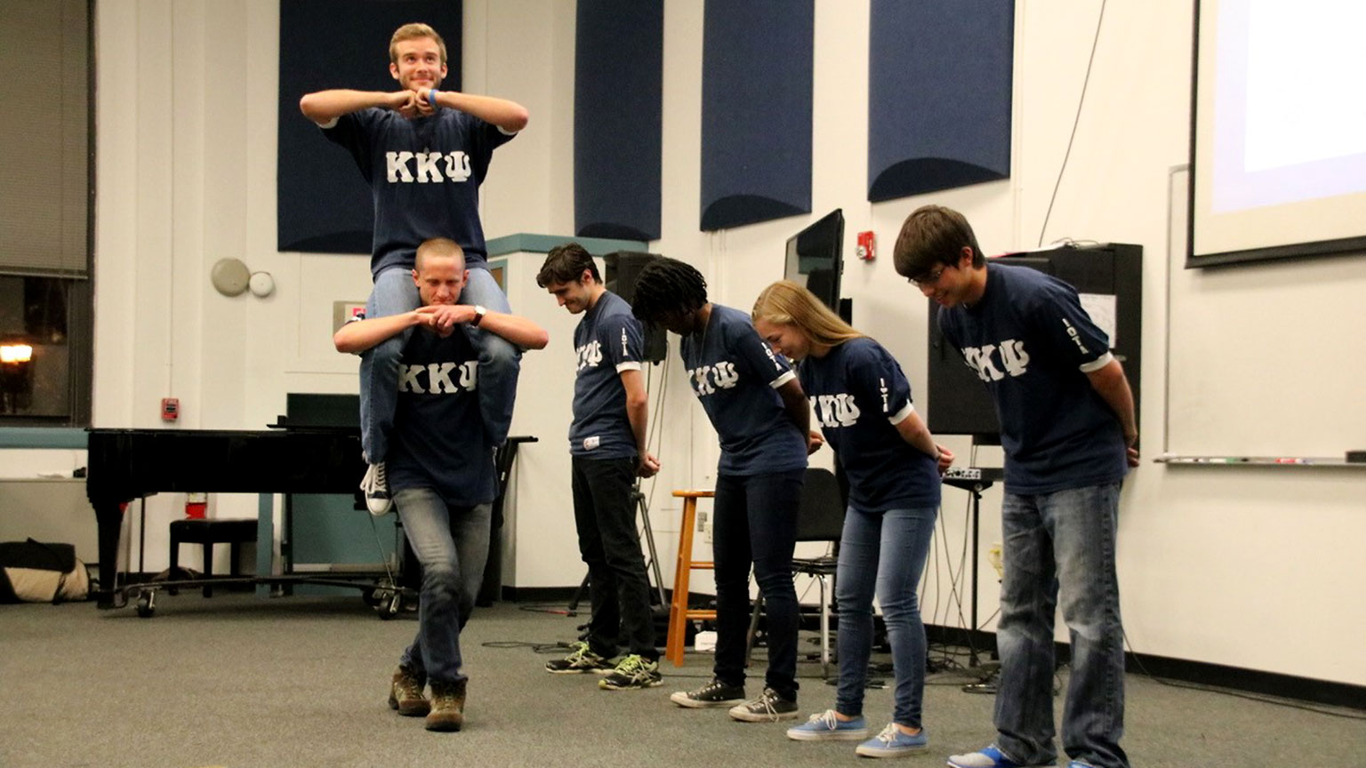 A member of the Kappa Kappa Psi marching band fraternity rides on a fraternity brother's shoulders while four other fraternity members line up and bow as the first two walk by. All people in this photo are wearing shirts with Greek letters for Kappa Kappa Psi on them.
