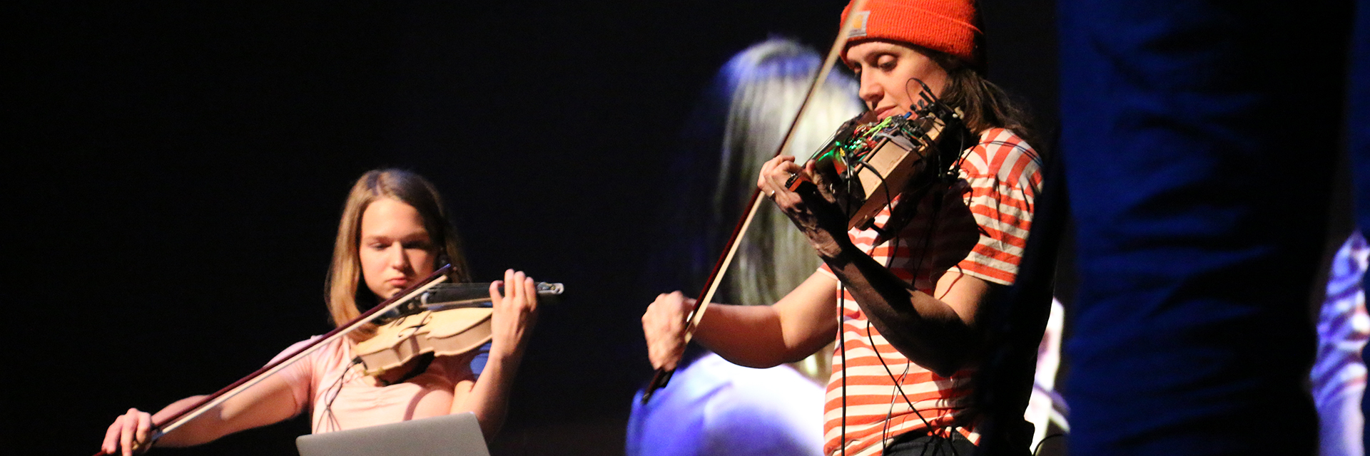 A girl playing a violin on stage next to a woman playing a modified violin with a circuit board on top.
