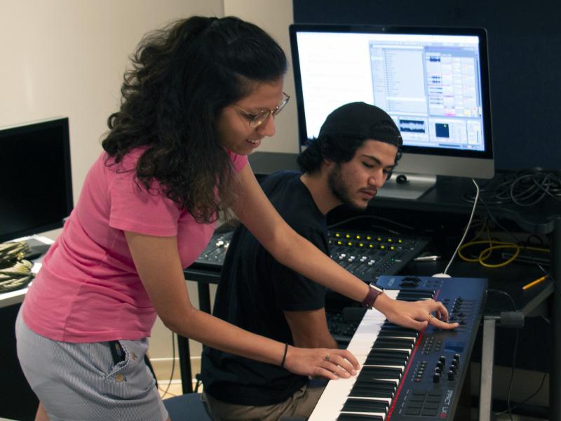 A female student plays the keyboard while a male student watches to record her in a studio.
